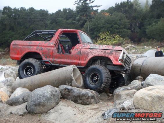 Red 1978 Ford Bronco with roll cage, off-roading over rocks and concrete pipes