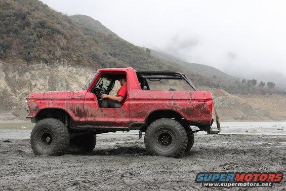 Red 1978 Ford Bronco with large tires driving through mud, driver visible