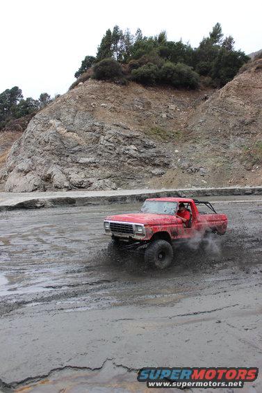 Red off-road truck with a roll cage driving through mud, front grille visible