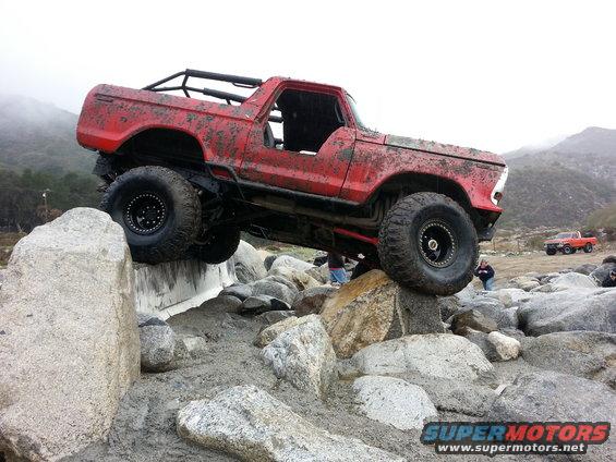 Red 1978 Ford Bronco with a roll cage, covered in mud, climbing over rocks