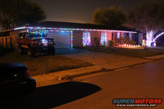 Red SUV with Christmas lights on roof and grille parked in driveway of house also decorated with lights