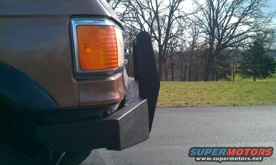 Front corner of a brown SUV with a black push bar and amber turn signal. Trees and a grassy hill in background