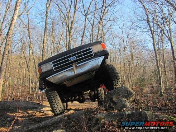 Ford truck with large tires navigating rocks on a wooded trail. Another vehicle is visible behind it