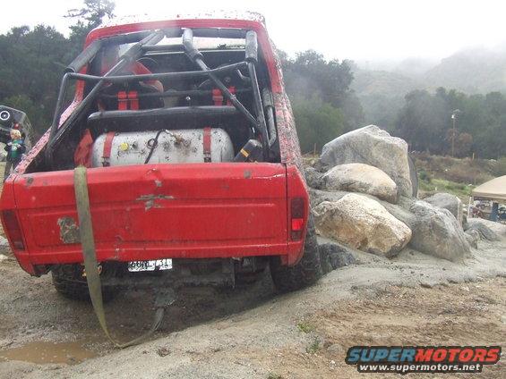 red pickup truck with roll cage and external fuel tank, going over rocks