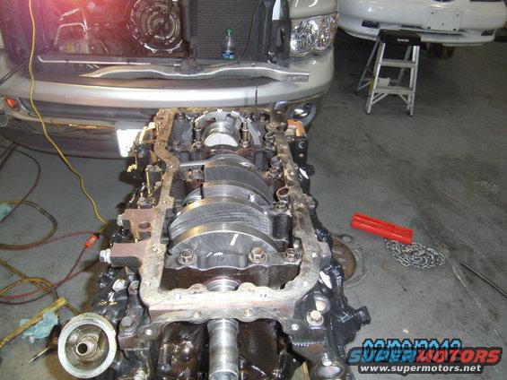 Engine block with crankshaft exposed, in front of a silver truck. Tools and a ladder are visible