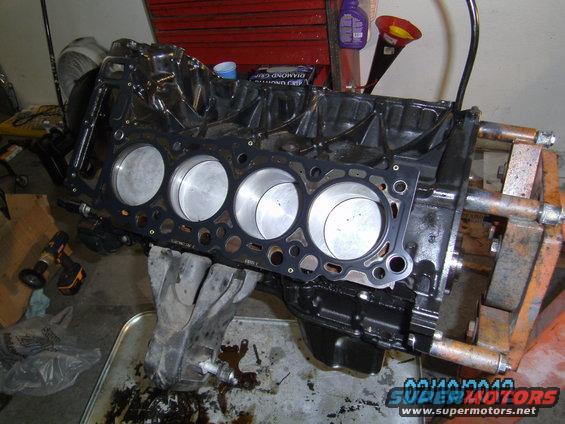 Engine block with four cylinders, pistons, and a gasket installed. A drill and shop rags are visible nearby