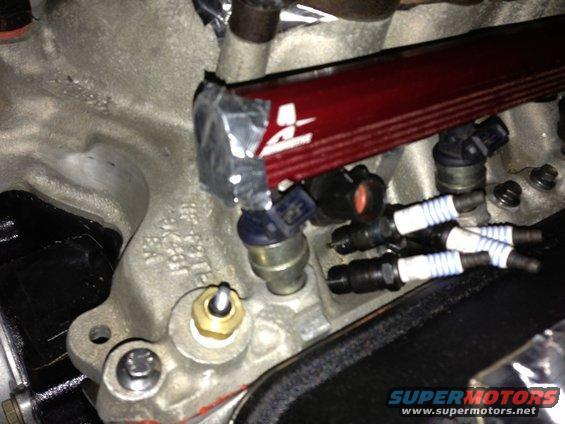 Engine manifold with red fuel rail, blue-capped injectors, and spark plugs. Some tape is visible on the fuel rail