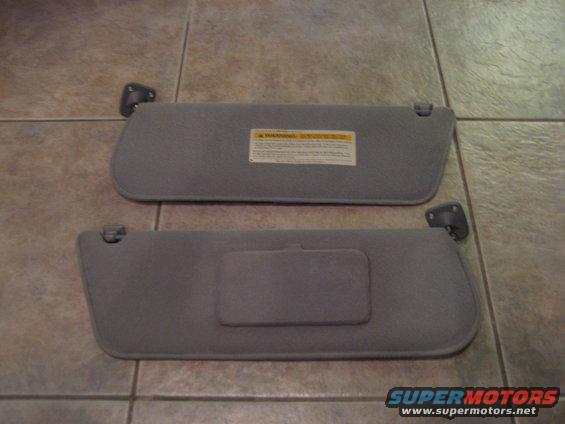 Two gray car sun visors on a tiled floor. One has a yellow warning label