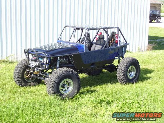 Custom buggy with roll cage, big tires, and exposed engine sits on grass. Another Jeep is parked nearby