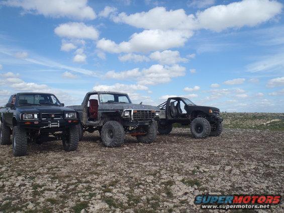 Three modified trucks parked on rocky ground under a cloudy sky. The middle truck appears to be a 1979 Ford Bronco