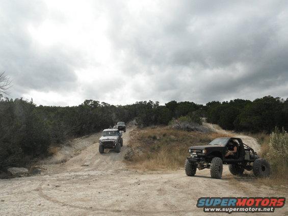 Several off-road vehicles drive up a dirt path with trees and cloudy sky