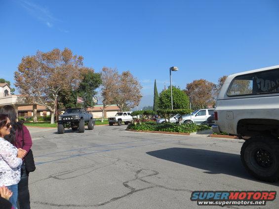 Several lifted trucks and SUVs parked in a lot under a clear blue sky. A white SUV is in the foreground