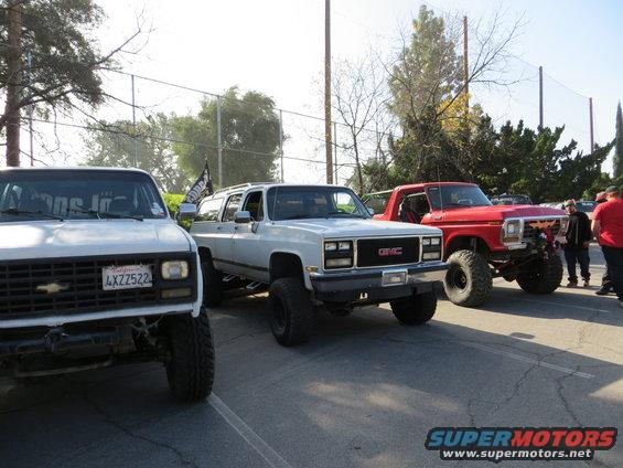 Three lifted SUVs parked in a lot, a white Chevrolet, a white GMC, and a red Ford