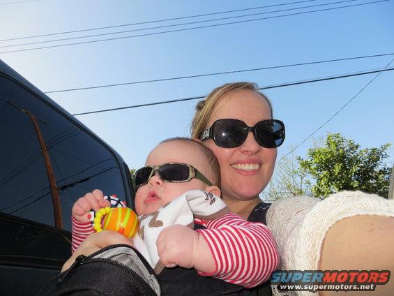 Woman holding a baby, both wearing sunglasses. They are next to a dark vehicle