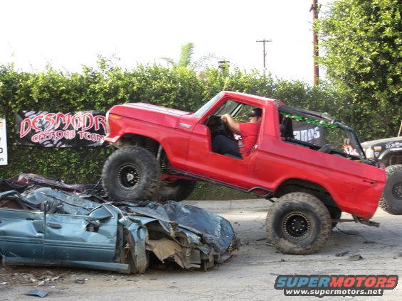 Red 1978 Ford Bronco driving over a crushed car. Banner in background says 'DESMODR OFFROAD TEAM'