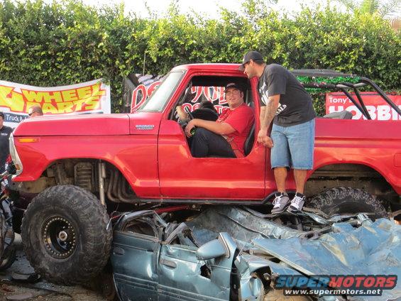 Red 1978 Ford Bronco lifted high, crushing a car underneath. Two men are in and around the Bronco