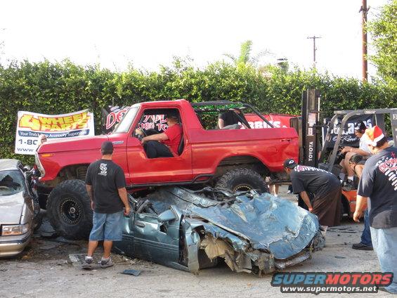 Red 1978 Ford Bronco crushing a blue car, lifted on large tires