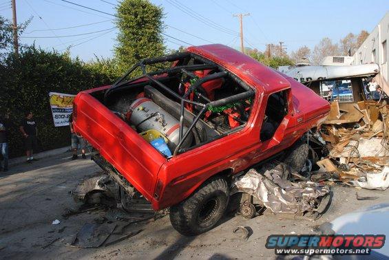 red off-road vehicle with roll cage, crushed under debris, next to a damaged bus