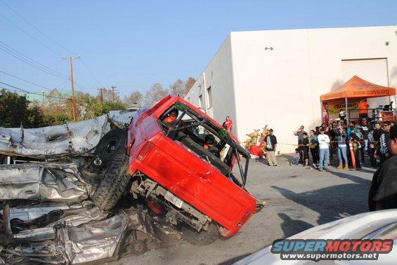 Red off-road vehicle on its side, crushed cars, crowd watching