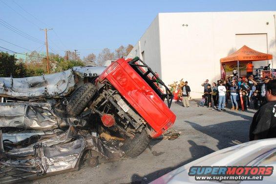 Red off-road vehicle flipped onto a pile of wrecked cars. Crowd watches in background