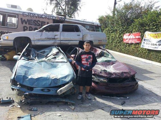 Boy standing between two heavily damaged cars. A white sedan is stacked on top of a bus in the background