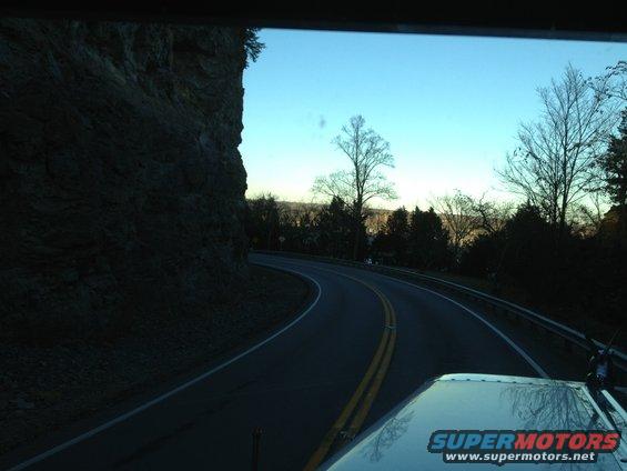 View from inside a vehicle looking at a winding road next to a cliff, with trees and sky in the background