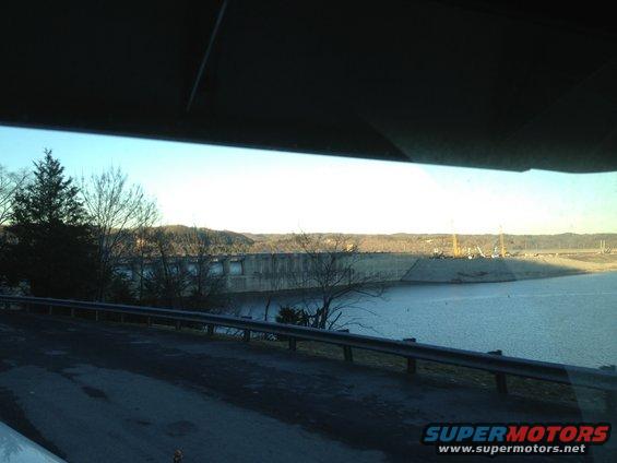 View of a large concrete dam and reservoir from a road, with trees and hills in the background