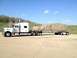 2003 Peterbilt 379 - A white semi-truck is parked on a concrete lot, hauling a large tarp-covered load on a flatbed trailer.