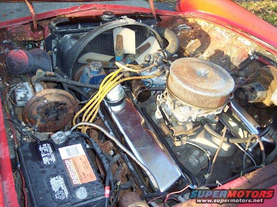 Engine bay showing V8 engine components including carburetor, valve covers, battery, and radiator