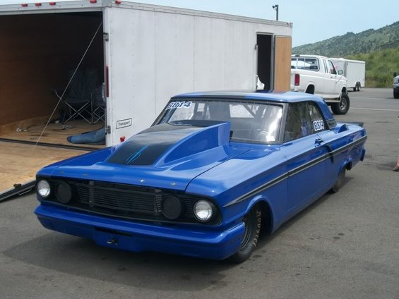 Blue 1964 Ford Falcon drag car with a large hood scoop and racing slicks parked next to a white trailer
