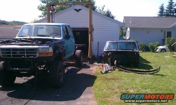 Light blue Ford F-Series truck chassis with front end removed, parked on asphalt. Another truck cab is in the yard