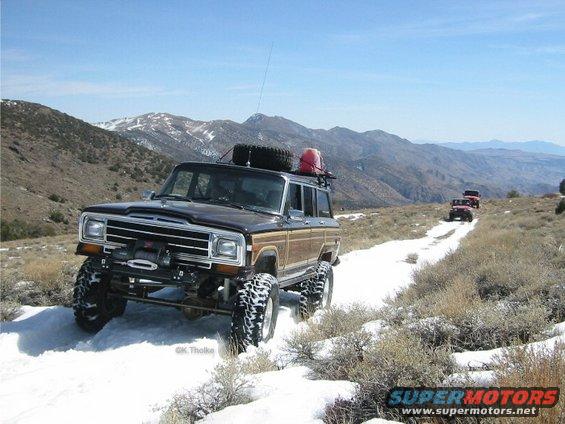 Brown Jeep Grand Wagoneer with wood paneling driving on a snowy mountain road, another red Jeep follows