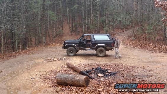 Black Ford Bronco parked on a dirt road in a wooded area, driver's door open, with a person standing beside it