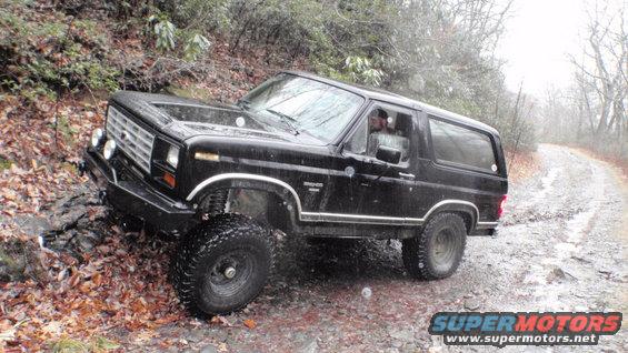 Black Ford Bronco with lifted suspension and off-road tires navigating a wet, wooded trail