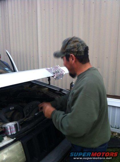 Man in camouflage hat works on car engine compartment; hood is open, showing engine parts and a roll of duct tape