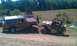 1987 Ford Bronco - A lifted Jeep tows a Ford Bronco through a grassy field with trees and a pond in the background.