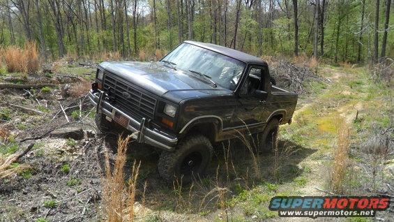 Black 1986 Ford Bronco driving on a dirt path in a wooded area