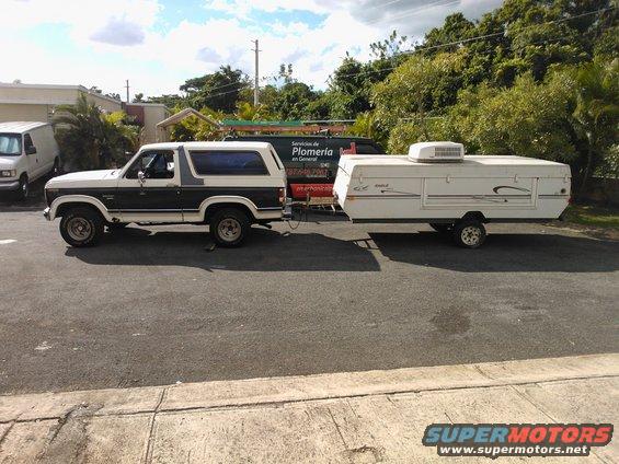 Side view of a white and brown 1984 Ford Bronco towing a white pop-up camper