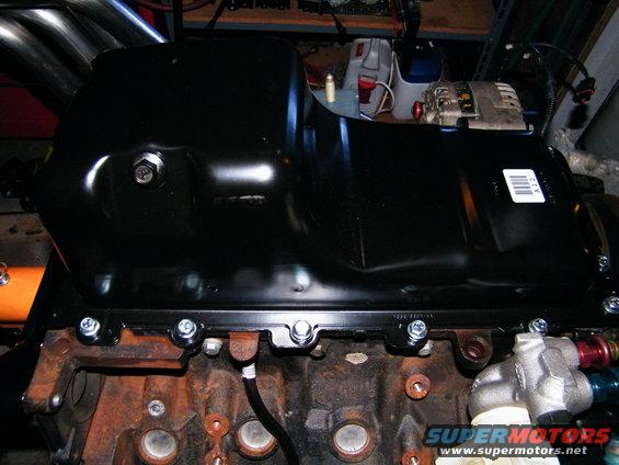 Black engine oil pan bolted to a rusty engine block with exhaust headers visible