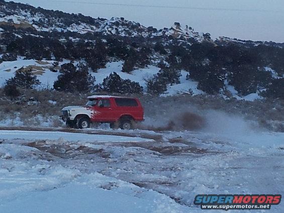 Red and white 1978 Ford Bronco driving through snow, kicking up dirt
