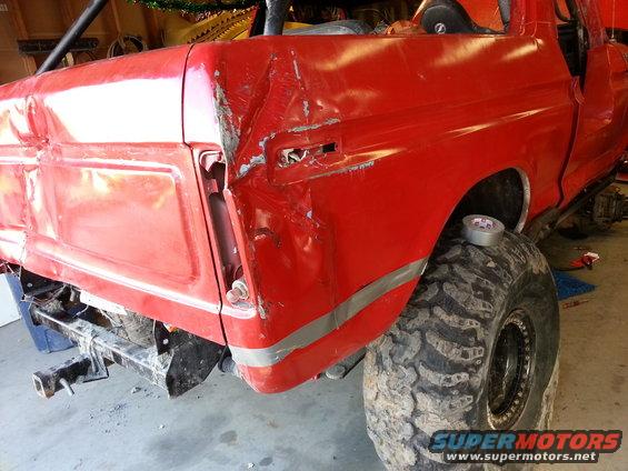 Red pickup truck rear with significant damage to the quarter panel and tailgate area; large off-road tire visible