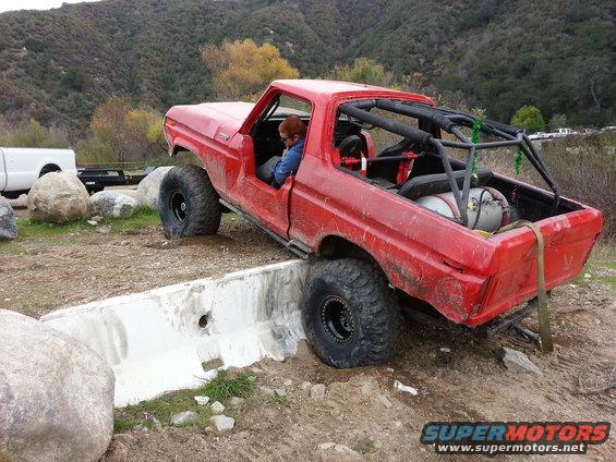 Red 1978 Ford Bronco with roll cage and large tires, tilted on a concrete barrier