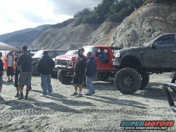 Red 1978 Ford Bronco with lifted suspension and large tires, parked on a dirt lot with other vehicles and people