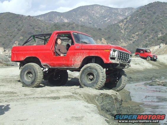 Red 1978 Ford Bronco with lifted suspension and large tires on a dirt trail, another red SUV in background