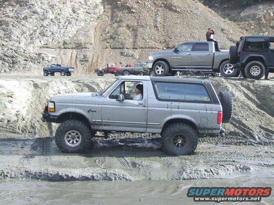 Silver Ford Bronco with lifted suspension and large tires driving through mud, other trucks visible in background