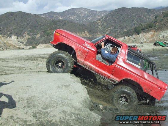 Red 1978 Ford Bronco off-roading, navigating a muddy ditch with a driver inside