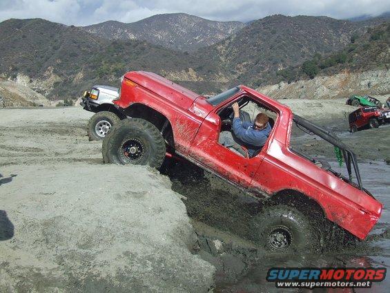 Red truck with roll cage going through mud, silver truck behind it