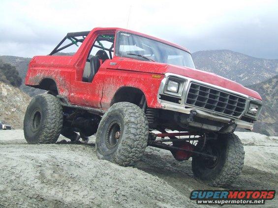 Red 1978 Ford Bronco with off-road modifications, including large tires and a roll cage, on a dirt hill