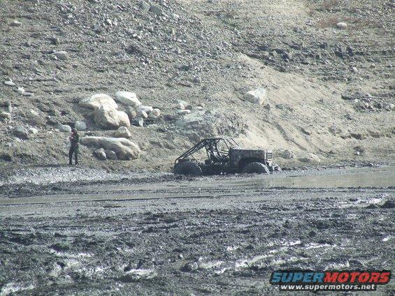 Mud-covered off-road vehicle with a roll cage stuck in thick mud, with a person standing on a rocky incline nearby