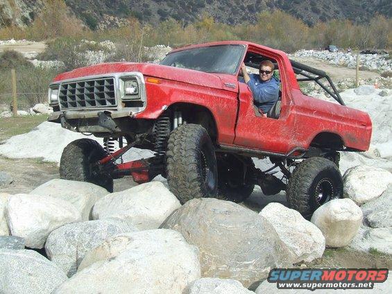 Red 1978 Ford Bronco with lifted suspension and large tires navigating a rocky terrain